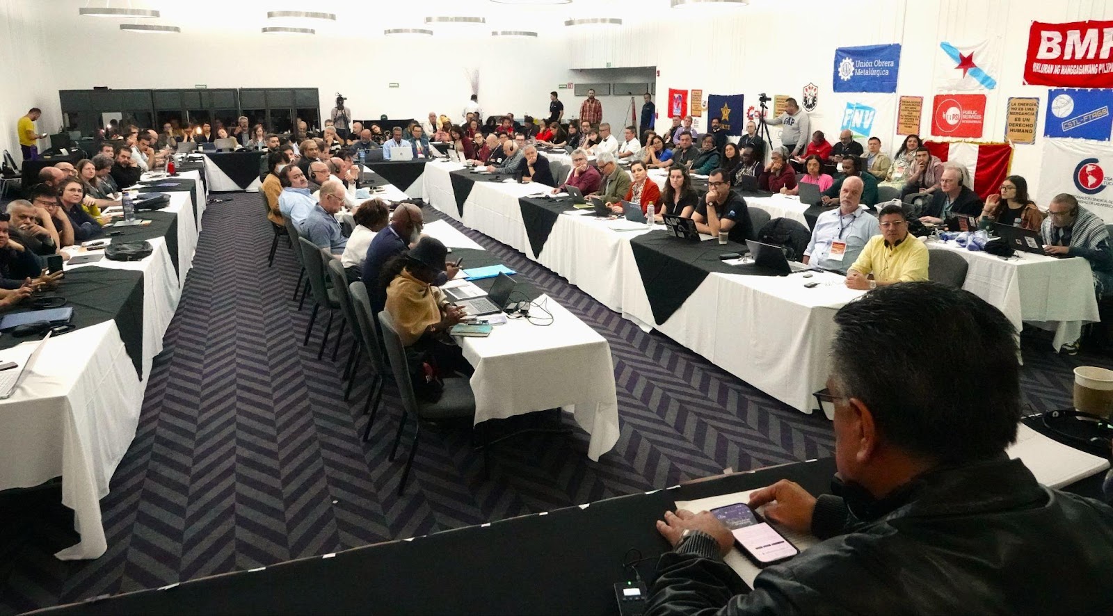 Wide view of a TUED meeting hall with delegates seated at long tables surrounded by union flags