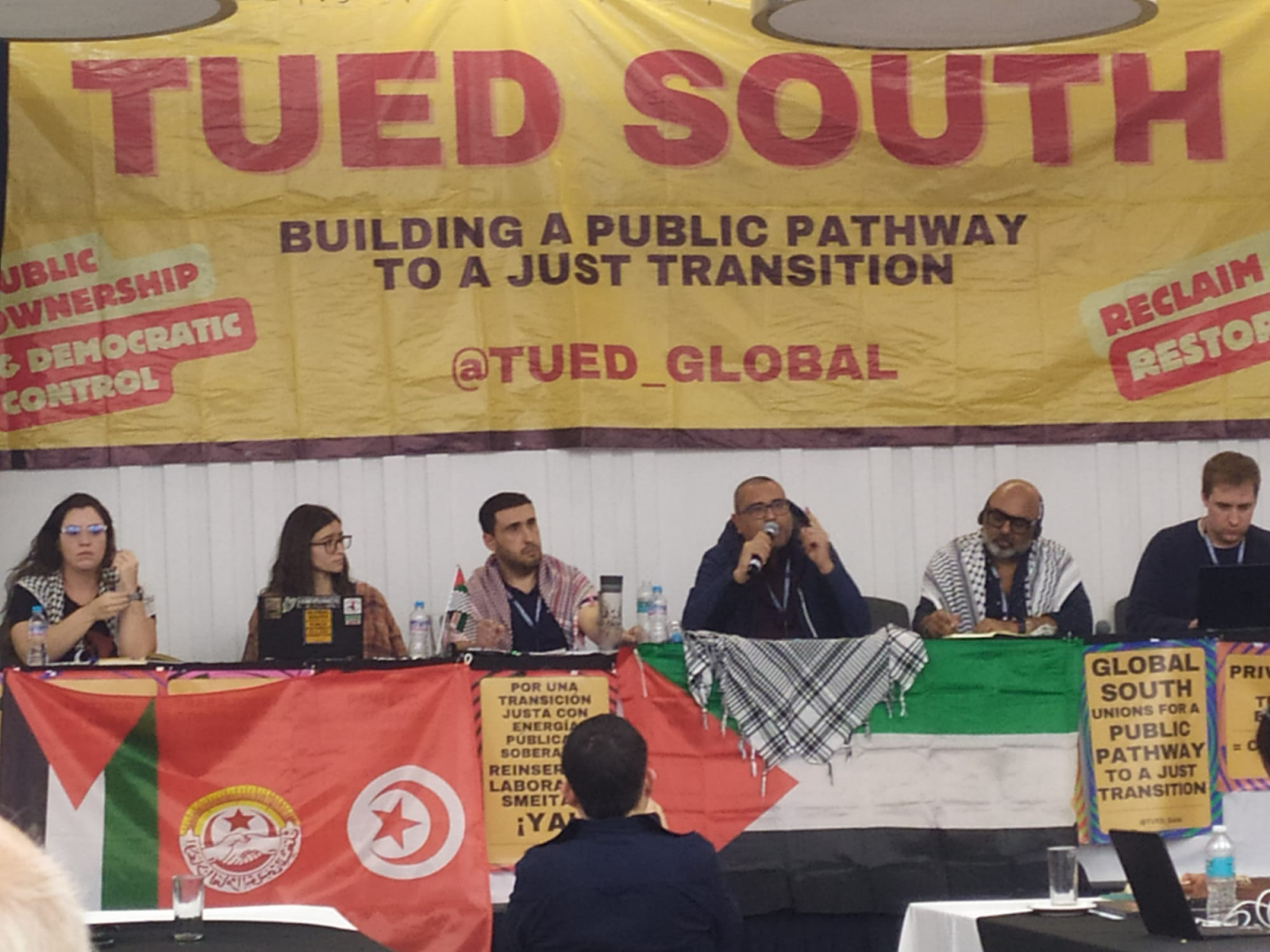 Panel of union leaders under the TUED South banner with flags of Palestine, Tunisia, and Algeria on the table